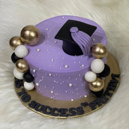 Purple graduation cake decorated with gold, black, and white edible spheres, featuring a graduation cap on top.