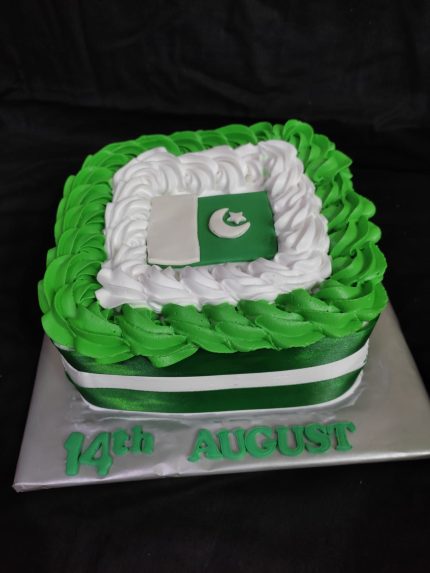 A square cake decorated with green and white frosting and a Pakistan flag on top, with '14th August' written at the base.